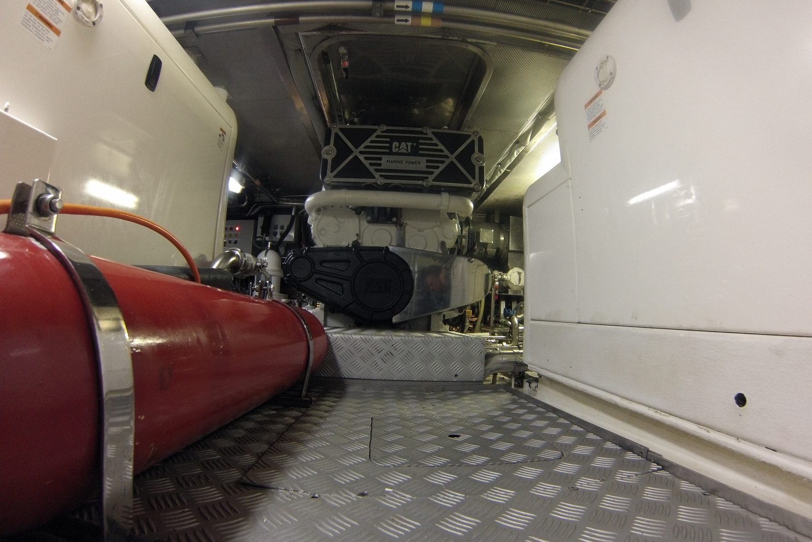 Engine room of a motor yacht: a marine diesel engine between white machinery enclosures, a red tank in the foreground and a diamond-plate deck