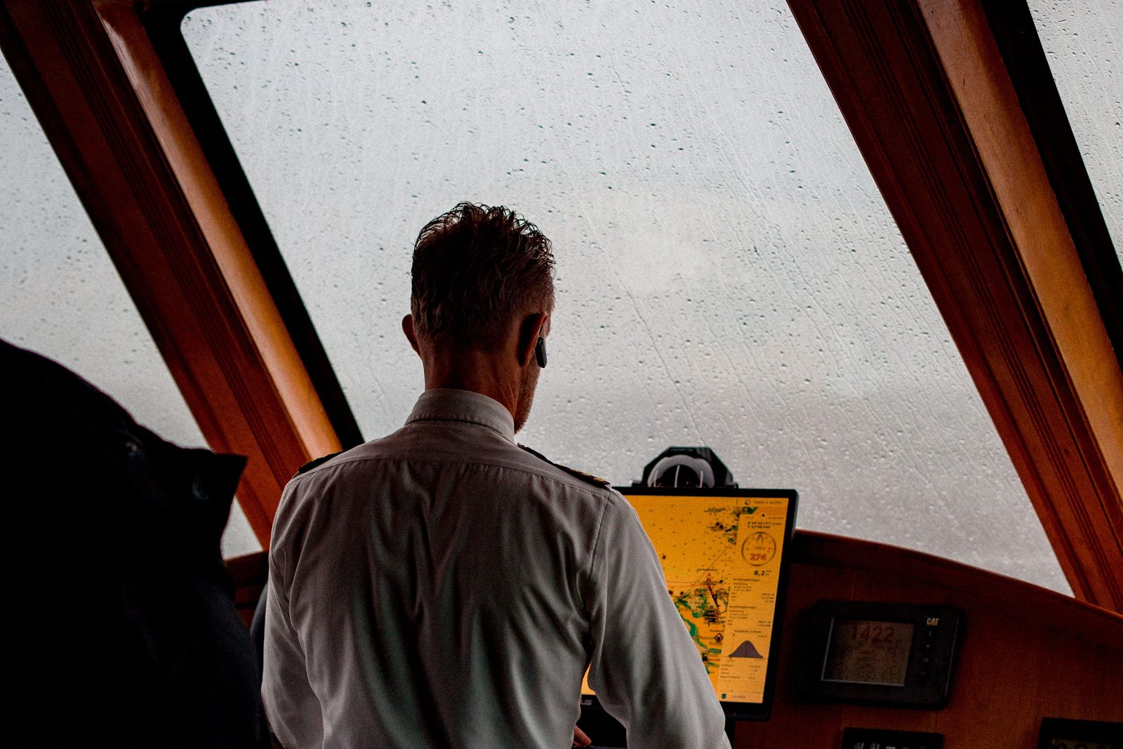 A yacht captain in a white uniform shirt, seen from behind, at the helm station beside a chartplotter on a rainy day