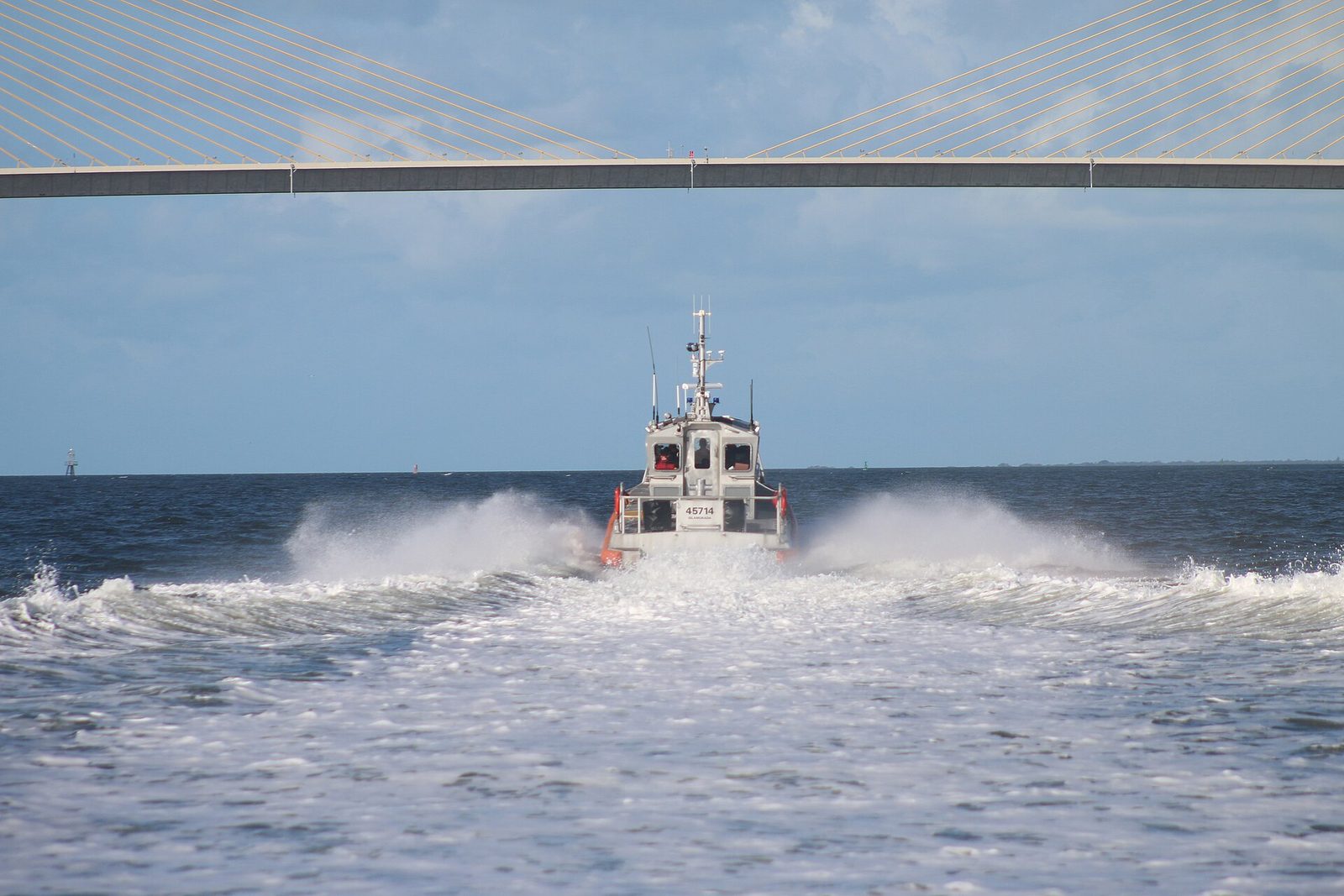 A U.S. Coast Guard response boat throws a wide white wake as it heads out beneath the cables of a long bridge on a bright day
